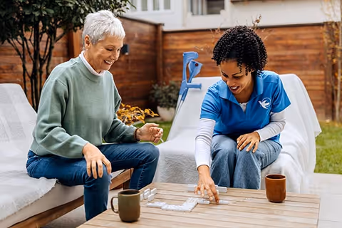 An elderly woman and a caregiver play dominoes on an outdoor patio.