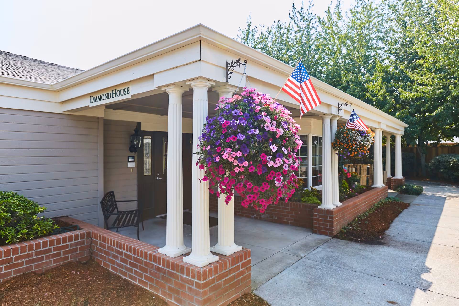 Exterior view of a building entrance with white columns and a brick base. Hanging flower baskets with pink, purple, and orange flowers are displayed, along with American flags. The building has a sign that reads 'Diamond House' above the entrance.