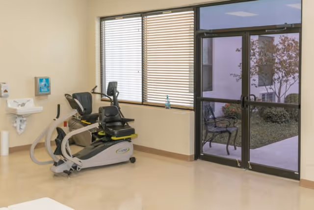 A small exercise room with a recumbent stationary bike positioned near a window with blinds. There is a wall-mounted sink with a soap dispenser and paper towel holder on the left side. A glass door on the right side leads outside to a patio area with a metal bench and some greenery.