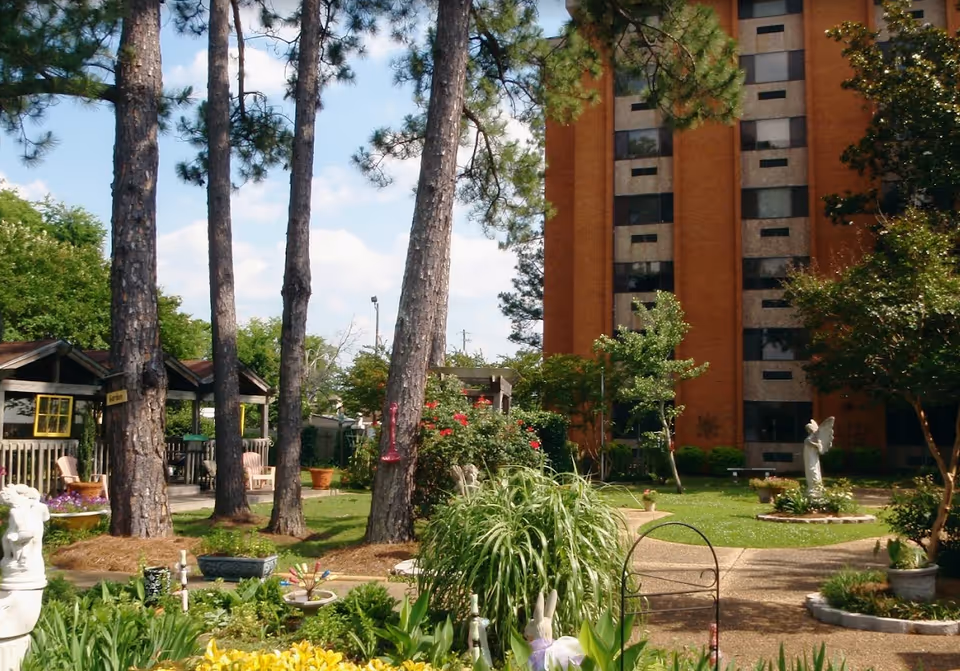 A garden area with various plants, flowers, and trees in front of a multi-story brick building. There are garden statues, a small gazebo with chairs, and paved walkways winding through the greenery under a partly cloudy sky.