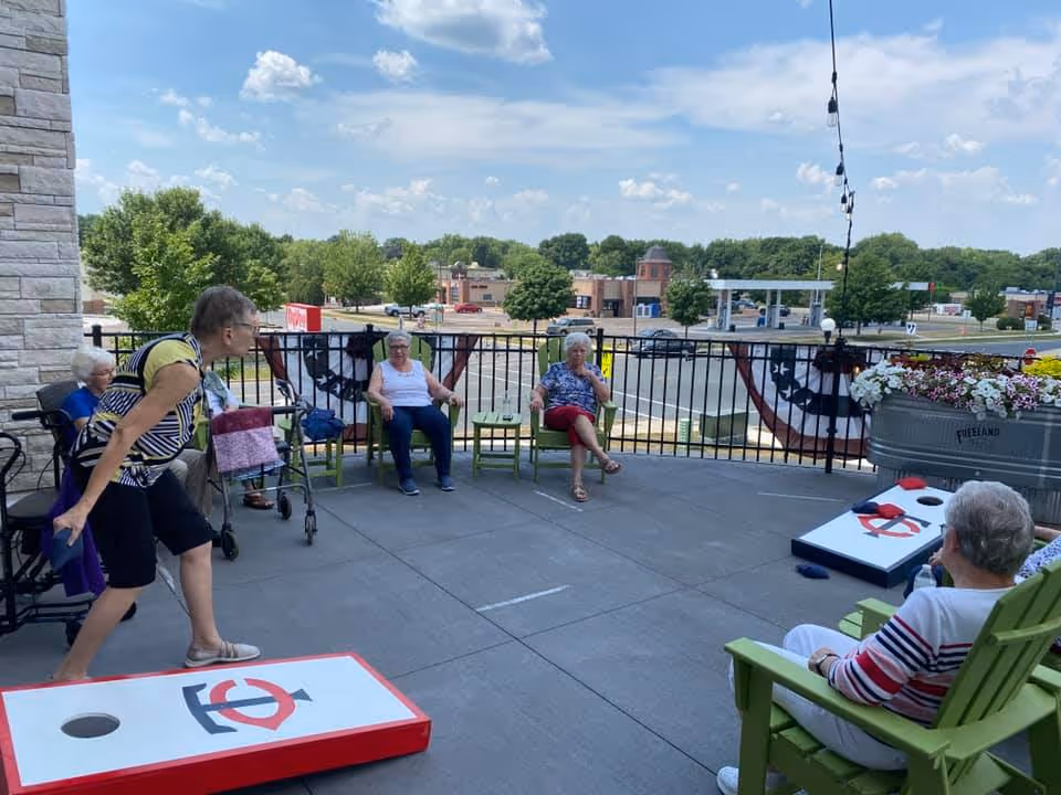 A group of elderly people sitting and standing on a patio playing cornhole on a sunny day. There are green chairs, a railing with patriotic bunting, and a planter with flowers. Trees and buildings are visible in the background under a partly cloudy sky.