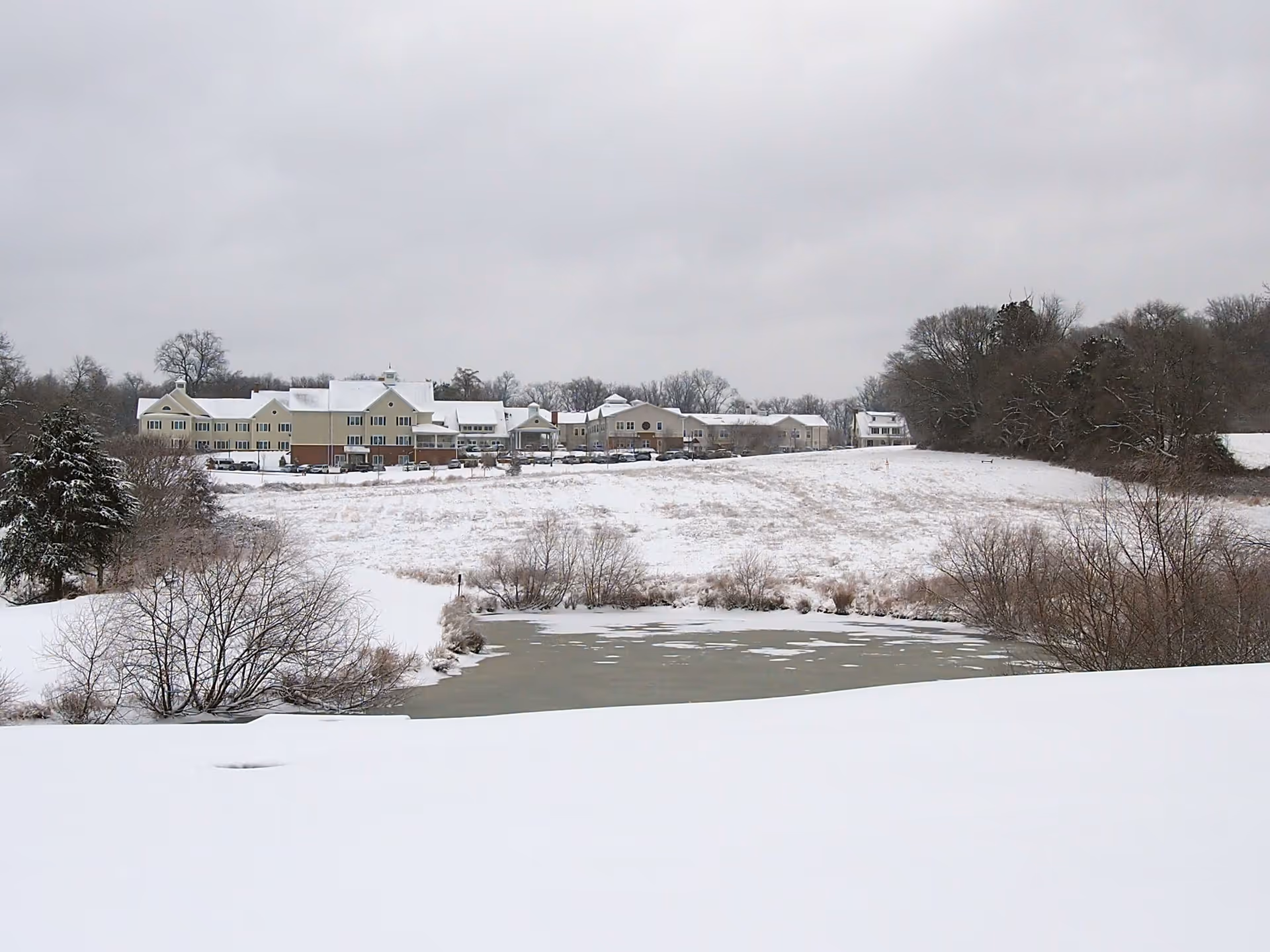 Snowy exterior of The Woods Assisted Living at Brooke Grove Retirement Village across a pond today.
