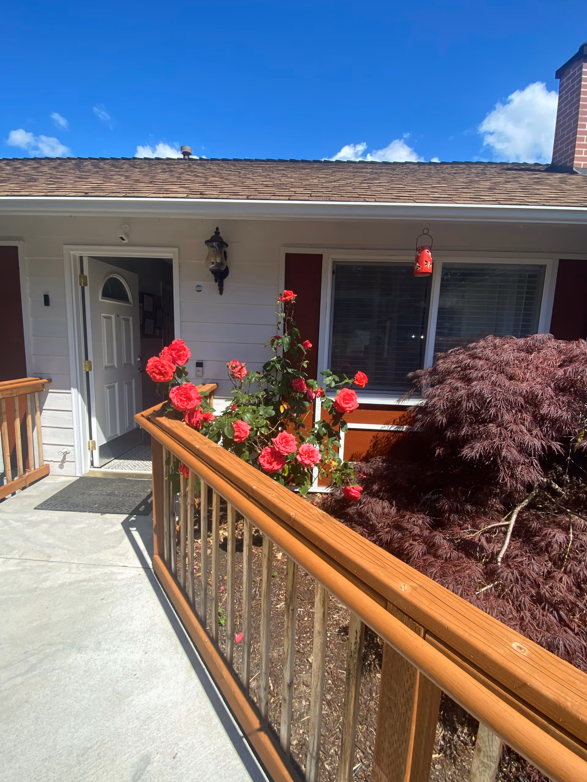 Exterior view of a single-story building with a wooden railing along a concrete walkway leading to an open white door. There are red roses and a bush with reddish leaves near the entrance under a clear blue sky.