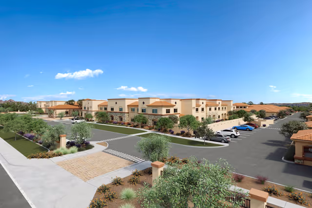 Exterior view of a low-rise Mediterranean-style senior living facility with landscaped grounds and a parking lot under a clear blue sky.