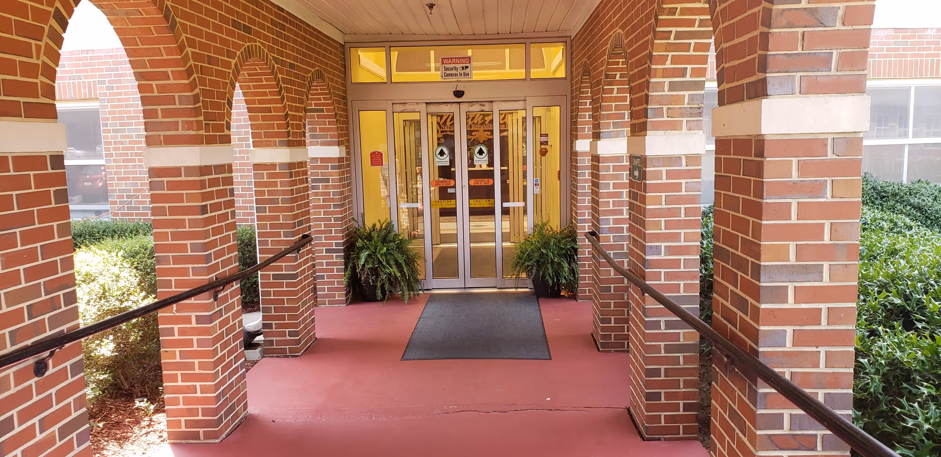 Covered brick-arched entrance with handrails and potted plants leading to glass double doors.
