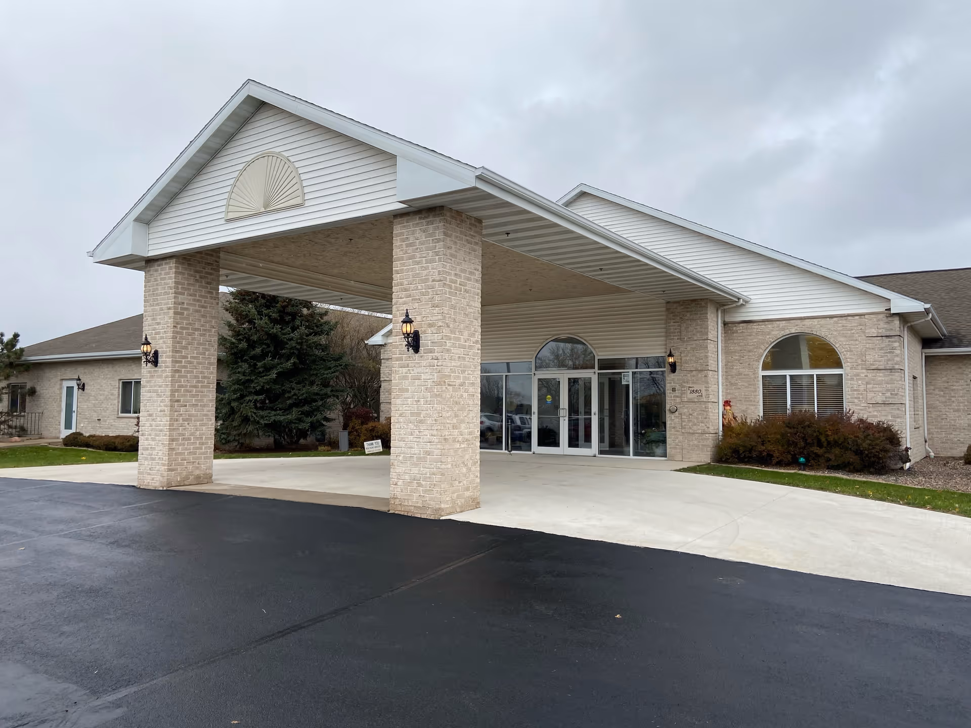 Exterior view of a senior living facility building with a covered entrance supported by brick pillars. The building has light-colored brick walls, large glass doors, and arched windows. The driveway and parking area are visible in front of the entrance under a cloudy sky.