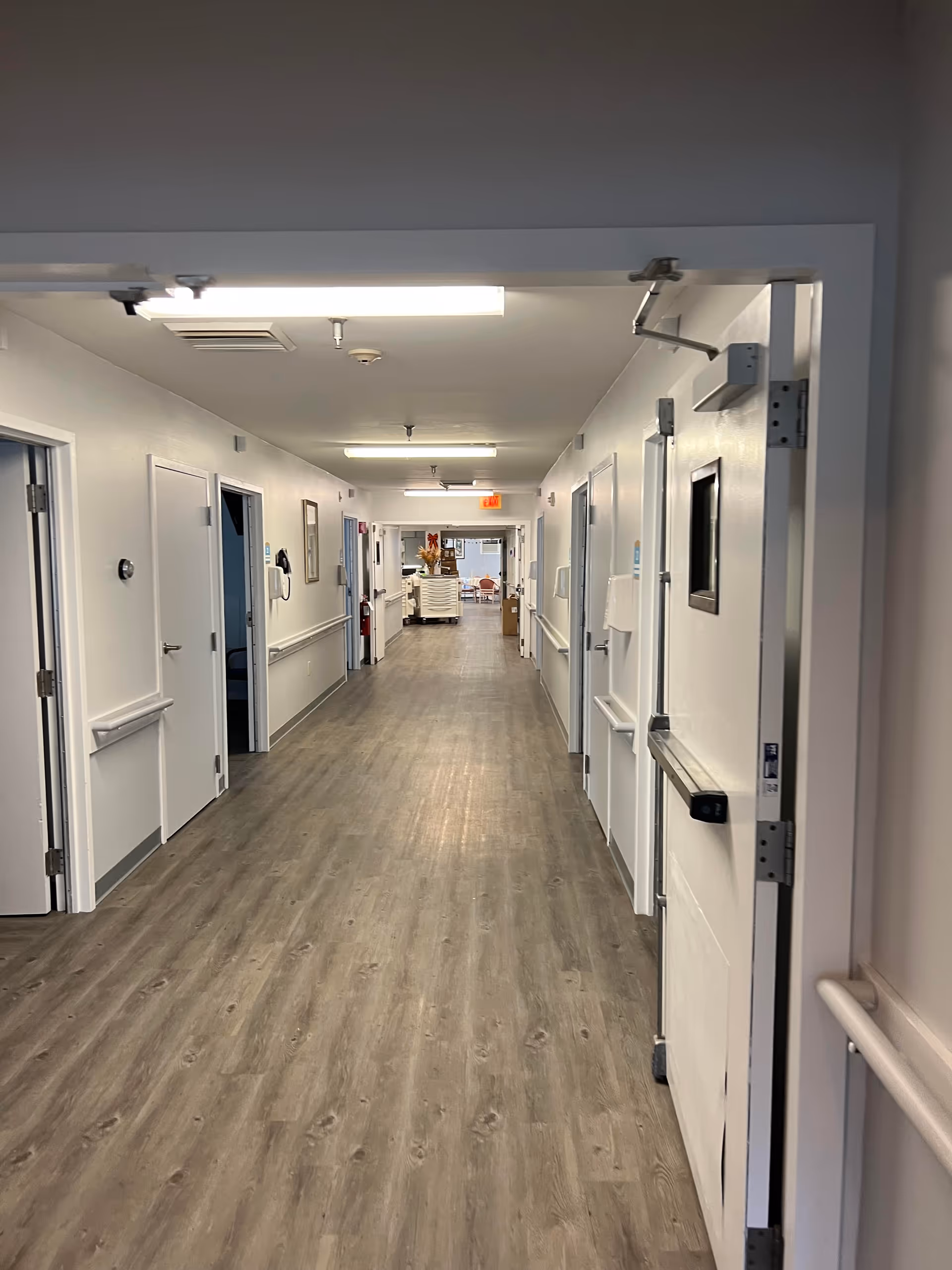 Long hospital-like hallway in a senior facility with doors and handrails on both sides.