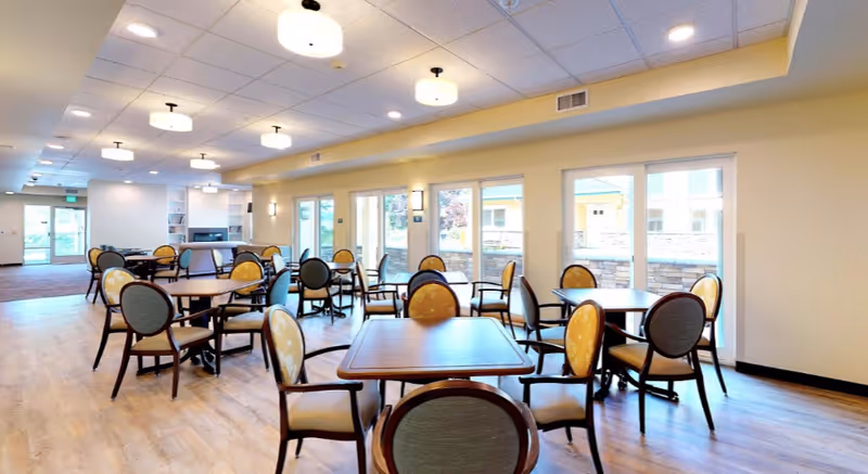 Bright communal dining room with multiple round tables and upholstered chairs on a wood floor and large windows.
