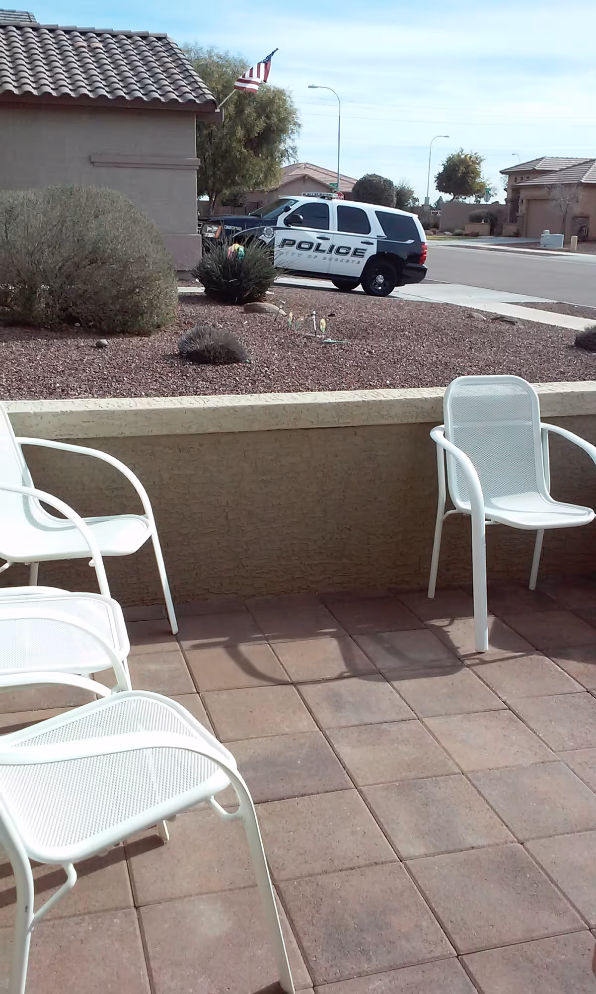 White metal patio chairs on a tiled front porch overlooking a gravel front yard and a parked police SUV near the curb.