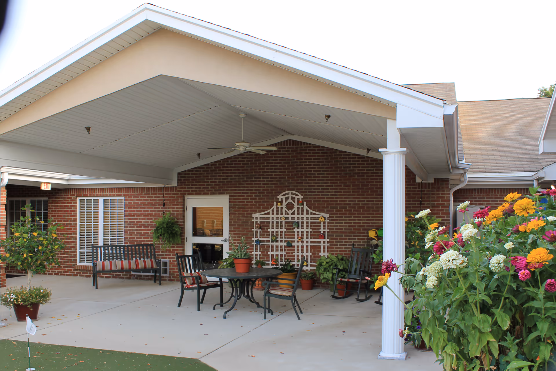 Covered brick patio with seating, table, potted plants, and colorful flowers.