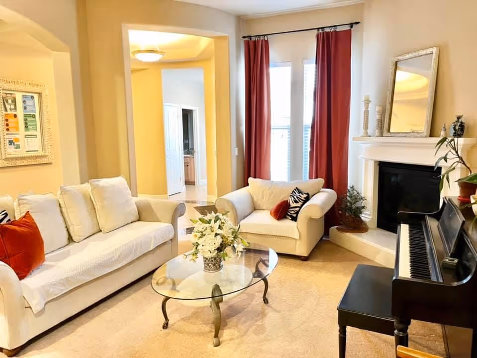 A cozy living room with a white sofa and armchair adorned with cushions, a glass coffee table with a floral arrangement, a black piano with a bench, a fireplace with decorative items on the mantel, red curtains covering tall windows, and a framed picture on the wall.