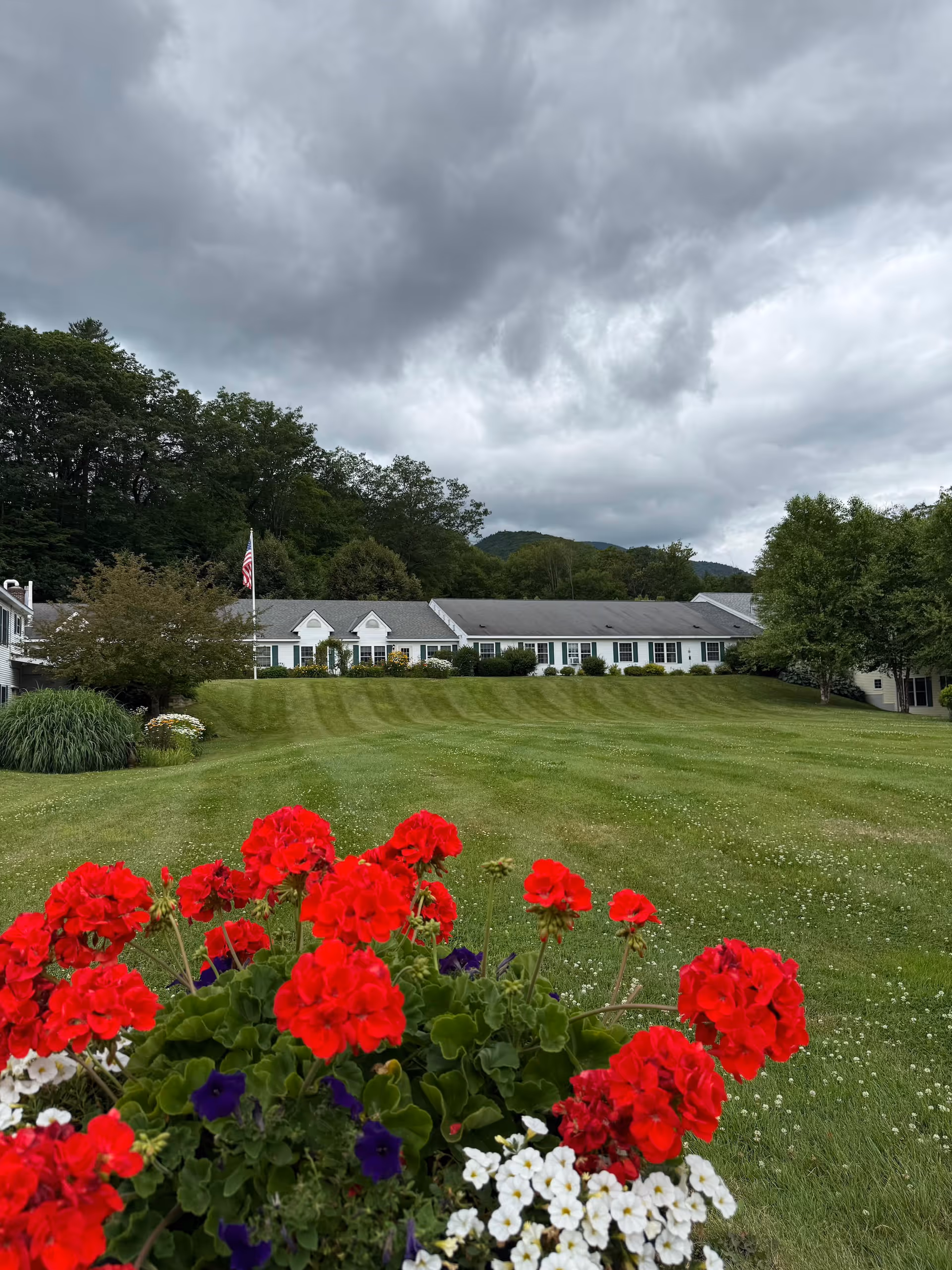 A wide view of the Village At Cedar Hill Inc facility showing a large green lawn with neatly trimmed grass and colorful red, purple, and white flowers in the foreground. The building is a long, single-story structure with white siding and multiple windows. Trees and hills are visible in the background under a cloudy sky.