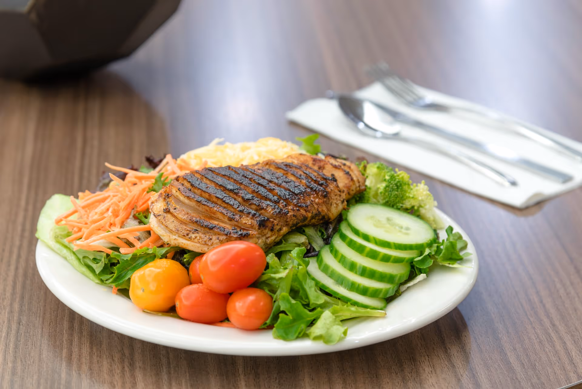 A plate of grilled chicken breast served on a bed of mixed greens, sliced cucumbers, cherry tomatoes, shredded carrots, broccoli, and shredded cheese on a wooden table with a fork and spoon on a napkin in the background.