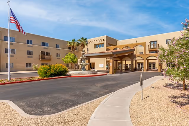 Front entrance of a three-story beige senior living building with a covered porte-cochere, fountain, flagpole, and desert landscaping under a blue sky.