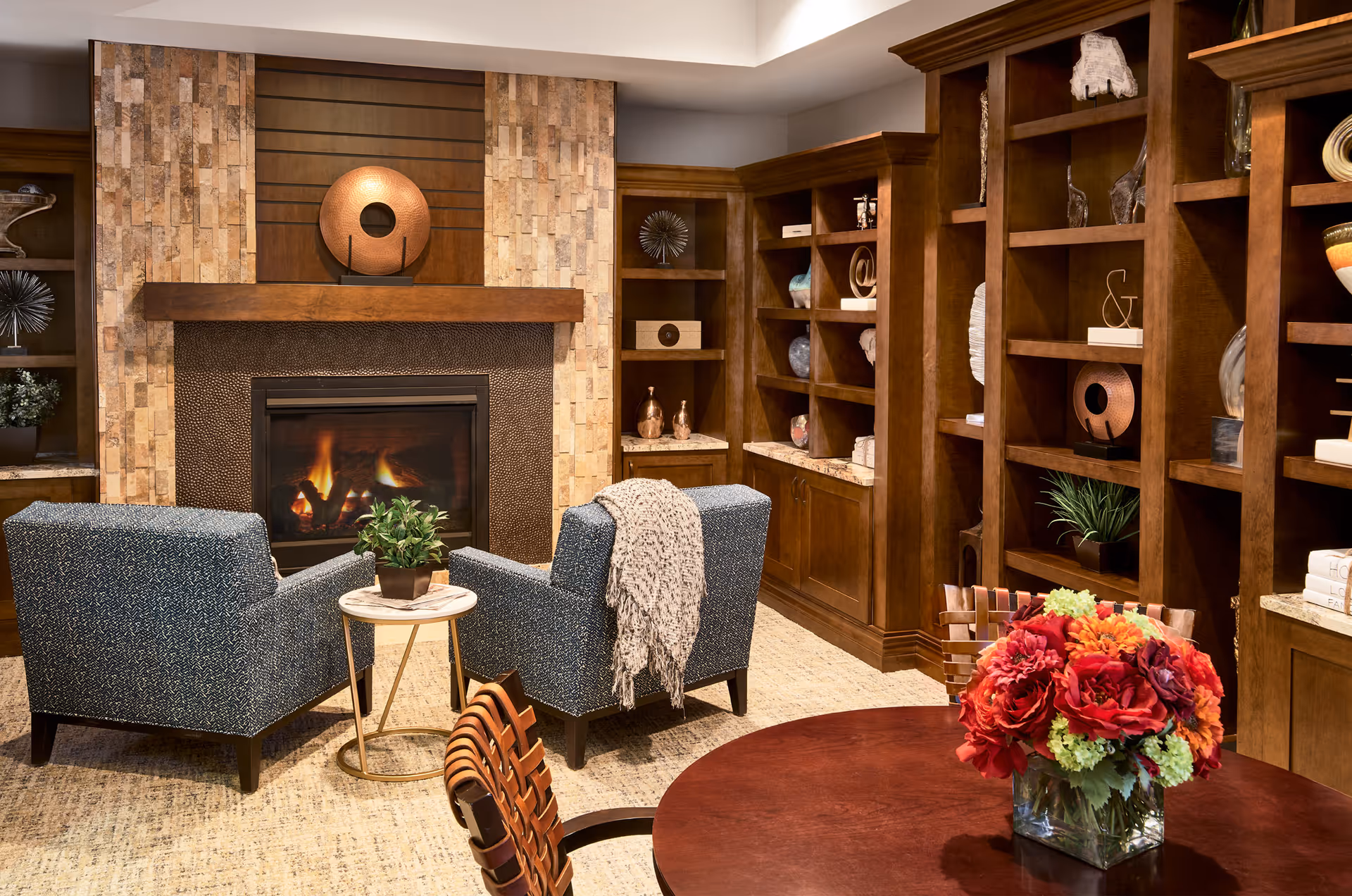 Cozy living room area with a lit fireplace surrounded by stone and wood paneling. Two blue upholstered armchairs face the fireplace with a small round side table between them holding a potted plant. Behind the chairs are built-in wooden shelves filled with decorative items. In the foreground, a round wooden table holds a vase with a colorful bouquet of red, orange, and green flowers.