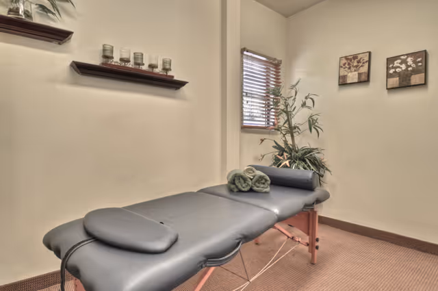 A small room with a padded massage table covered in gray upholstery, two rolled green towels placed on it, a window with wooden blinds, two framed floral pictures on the wall, a potted plant in the corner, and a wall shelf holding several candles.
