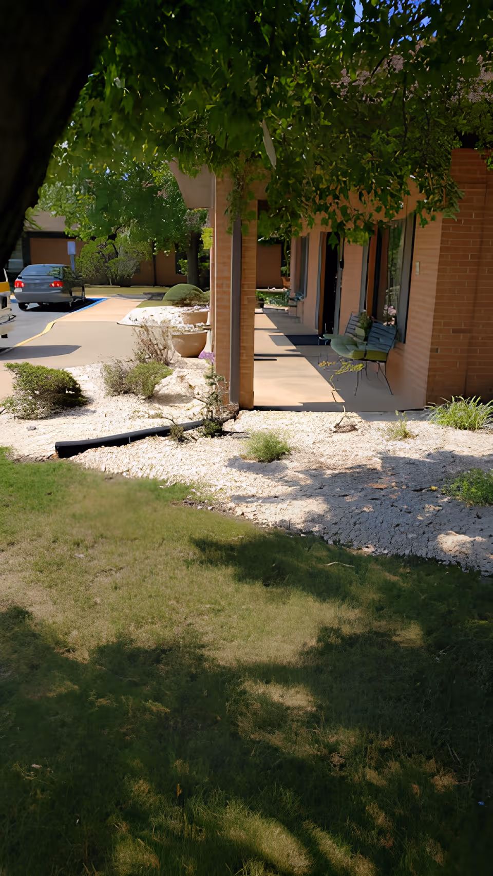 Covered walkway and entrance of a brick senior living facility with chairs, landscaping, and parked cars.
