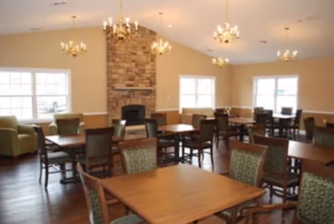 A spacious dining room with multiple wooden tables and chairs arranged neatly. The room features large windows allowing natural light to fill the space, a stone fireplace in the center of the far wall, and several chandeliers hanging from the ceiling. The walls are painted a warm beige color, and the floor is made of dark wood.