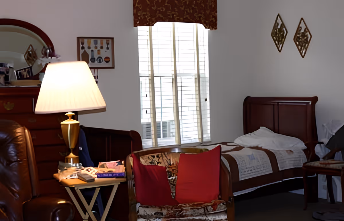 A cozy bedroom with a single bed covered with a quilt, a wooden dresser with a mirror, a table lamp on a small side table, a cushioned chair with red pillows, and a window with blinds and a valance.