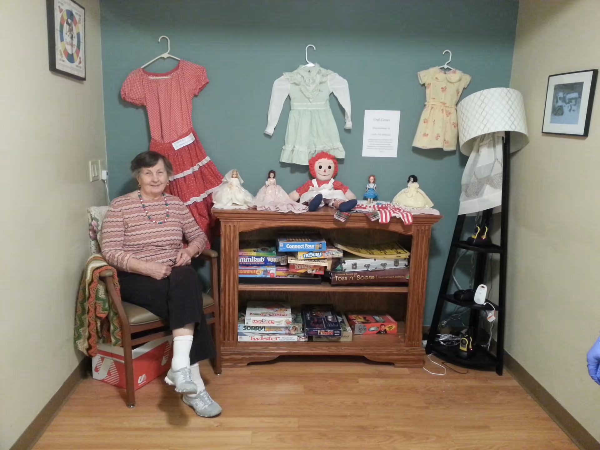 An elderly woman sitting on a chair next to a wooden shelf filled with board games and dolls. Three vintage dresses are hanging on the wall above the shelf, along with a sign labeled 'Craft Corner'. A floor lamp with a white shade and a black corner shelf with various items are to the right. The room has wooden flooring and light-colored walls.