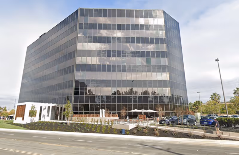 A tall glass front office building with reflective windows and a parking lot.