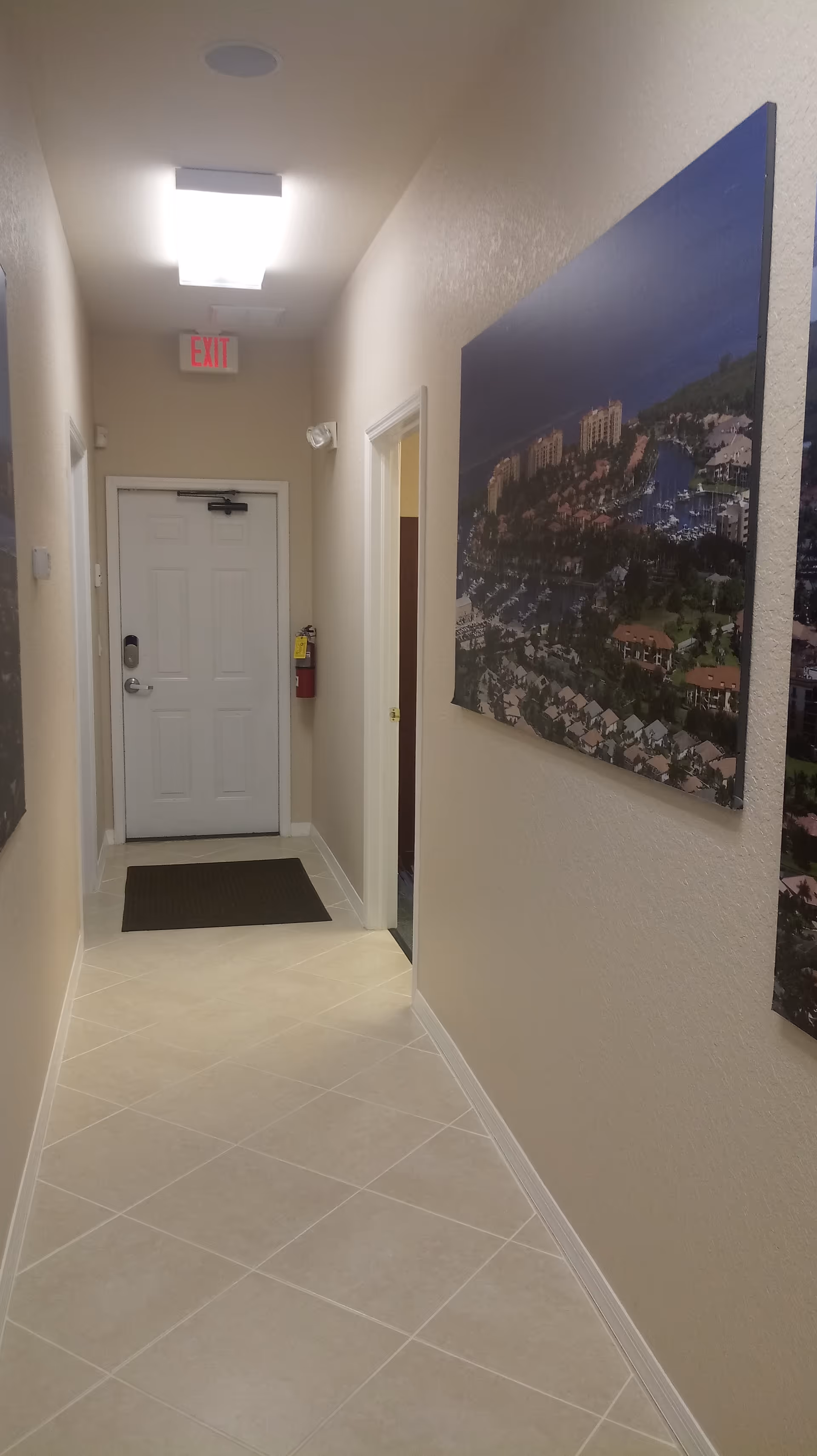 Narrow beige interior hallway with tiled floor, wall art, and an exit sign above a door.