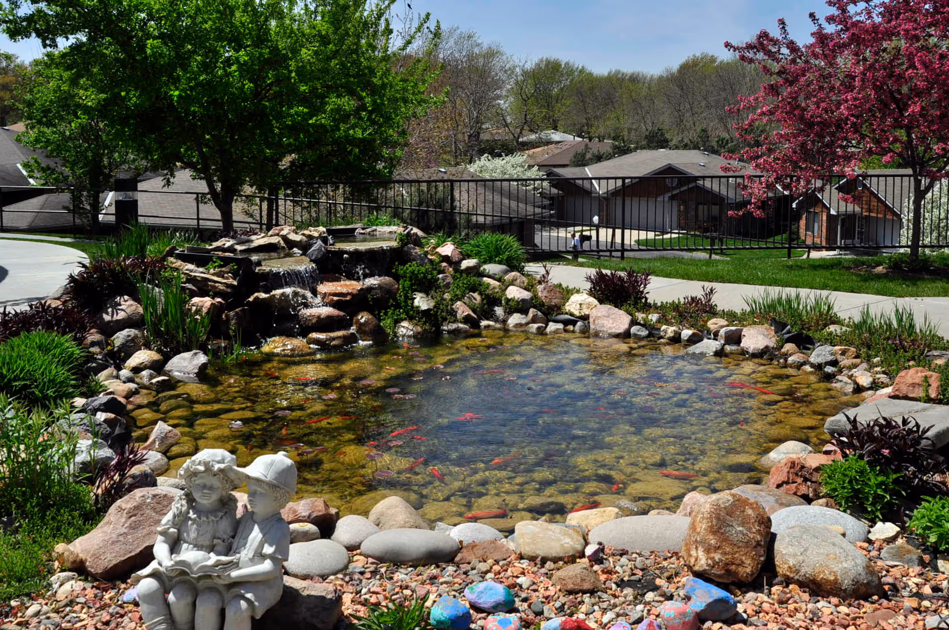Stone-lined pond with koi, surrounded by rocks, trees, and a statue in a landscaped garden.