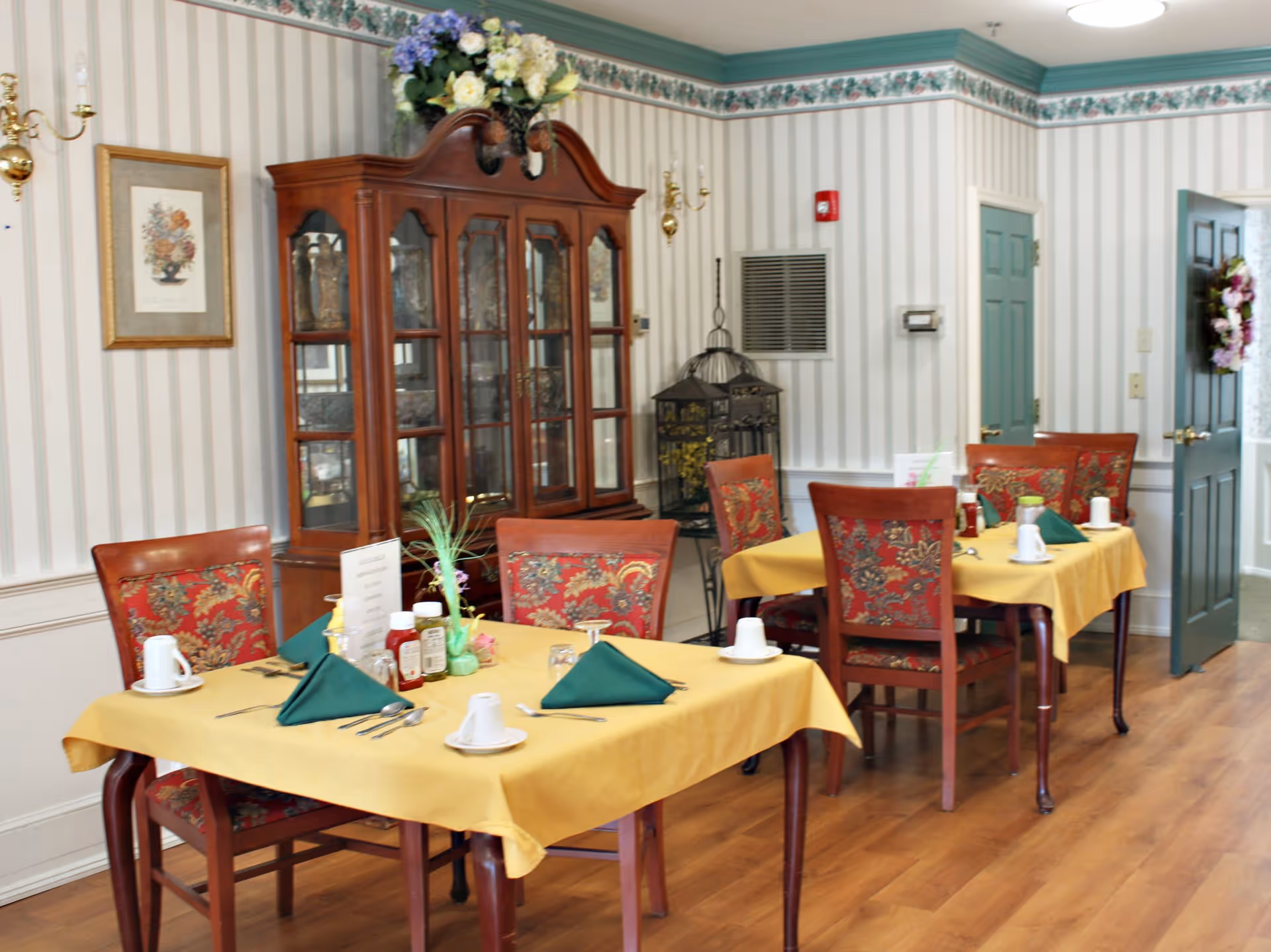 A dining room with two tables covered in yellow tablecloths, each set with green folded napkins, white cups, silverware, and condiments. The chairs have red and gold patterned upholstery. In the background, there is a wooden china cabinet with glass doors displaying decorative items and a floral arrangement on top. The walls have vertical striped wallpaper with a floral border near the ceiling, and there is a framed floral picture on the wall.