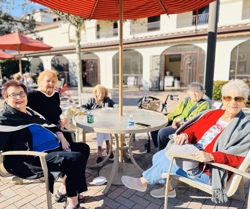 Five elderly women sitting around a round outdoor table with a red umbrella, enjoying a sunny day at a senior living facility. The background shows a building with arched doorways and windows.