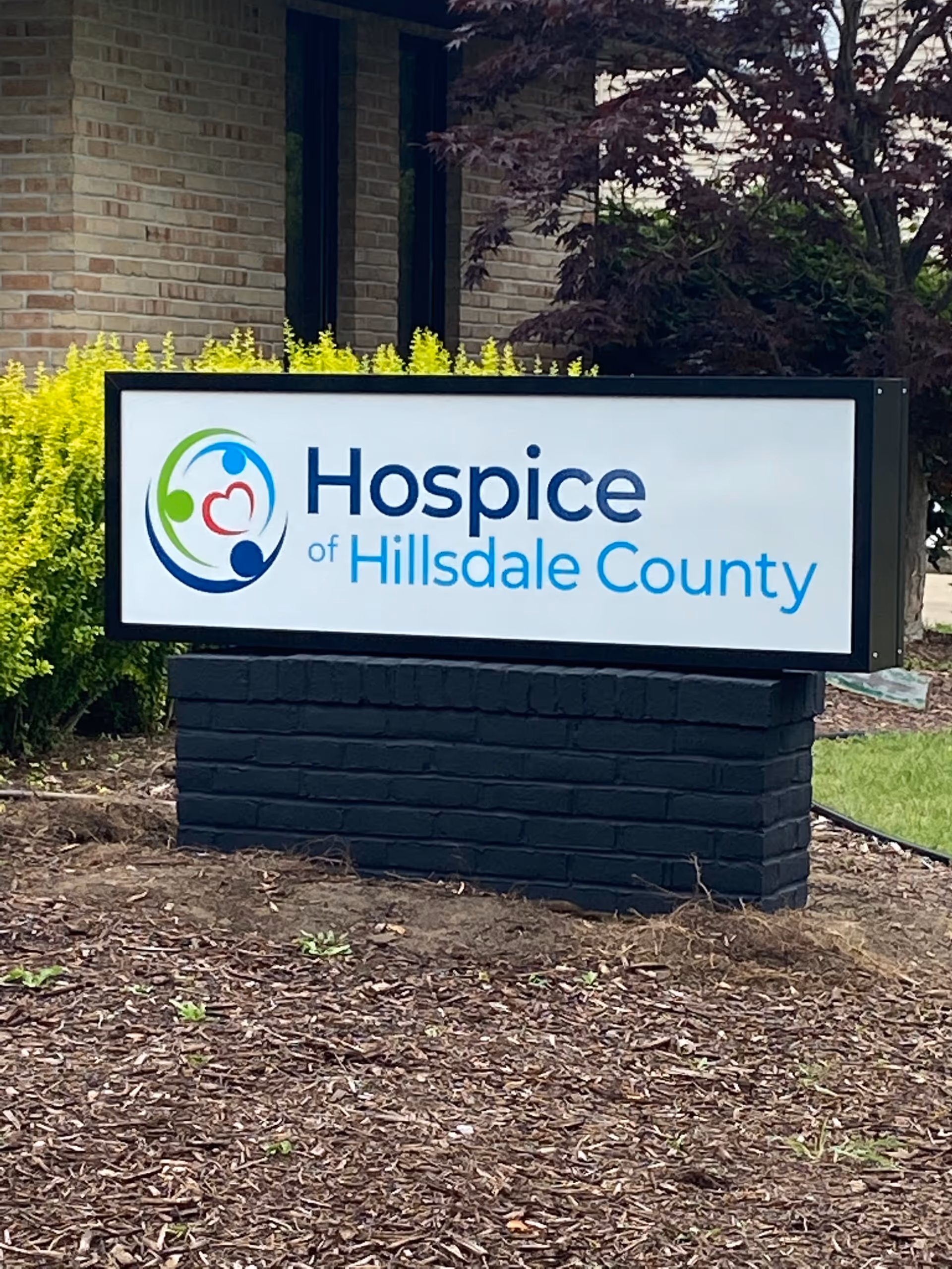 Outdoor sign reading Hospice of Hillsdale County with a colorful logo in a landscaped yard.