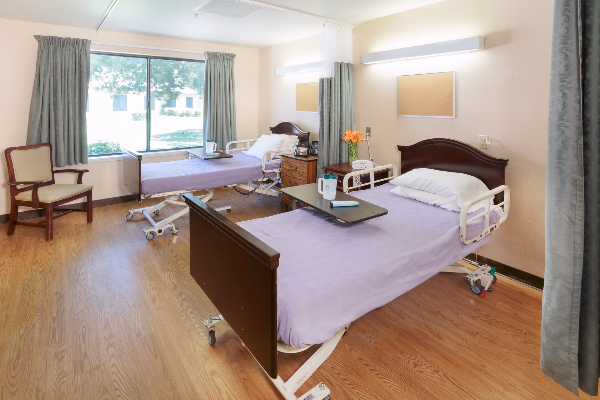 A bright and clean room with two hospital-style beds covered with light purple bedspreads. Each bed has a wooden headboard and white side rails. There is a small wooden nightstand between the beds with a telephone, a vase of orange flowers, and some framed photos. A large window with green curtains lets in natural light, and a wooden chair is placed near the window. The floor is wood laminate, and the walls are light-colored with two empty bulletin boards above the beds.
