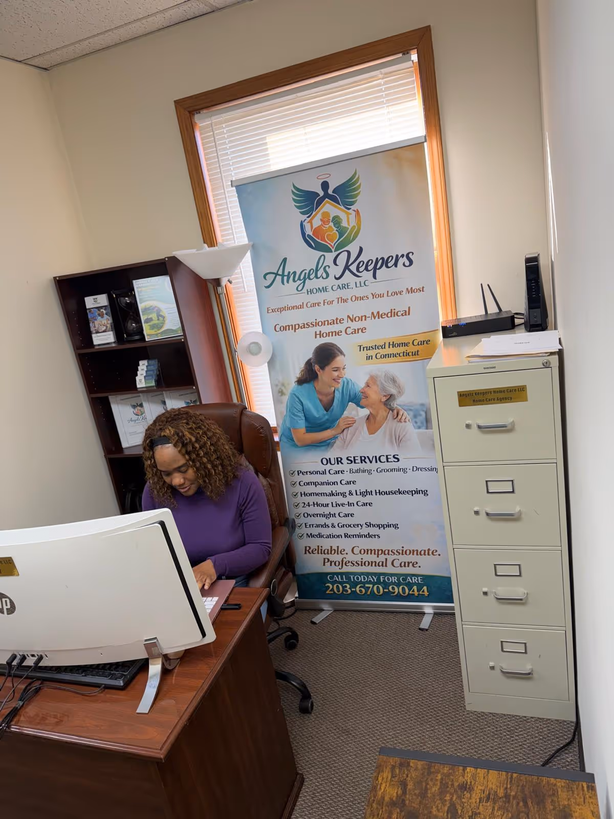 Office space with banner for Angels Keepers Home Care, a seated staff member at a desk.