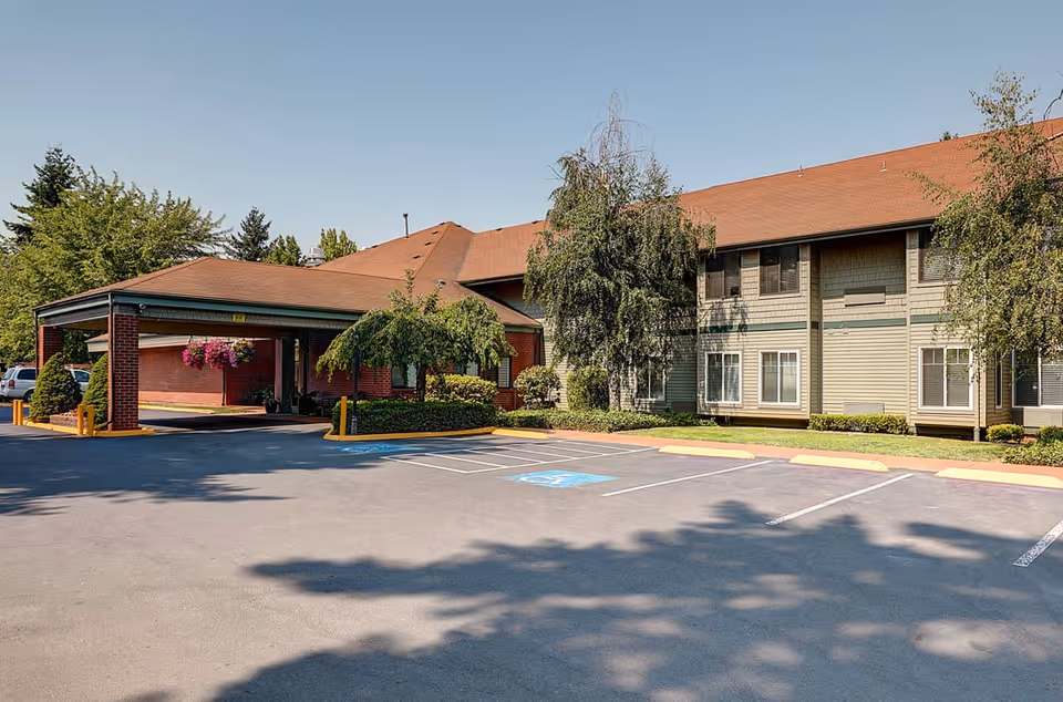 Exterior view of Firwood Gardens senior living facility showing a two-story building with a covered entrance, surrounded by trees and a parking lot with marked handicap spaces under a clear blue sky.