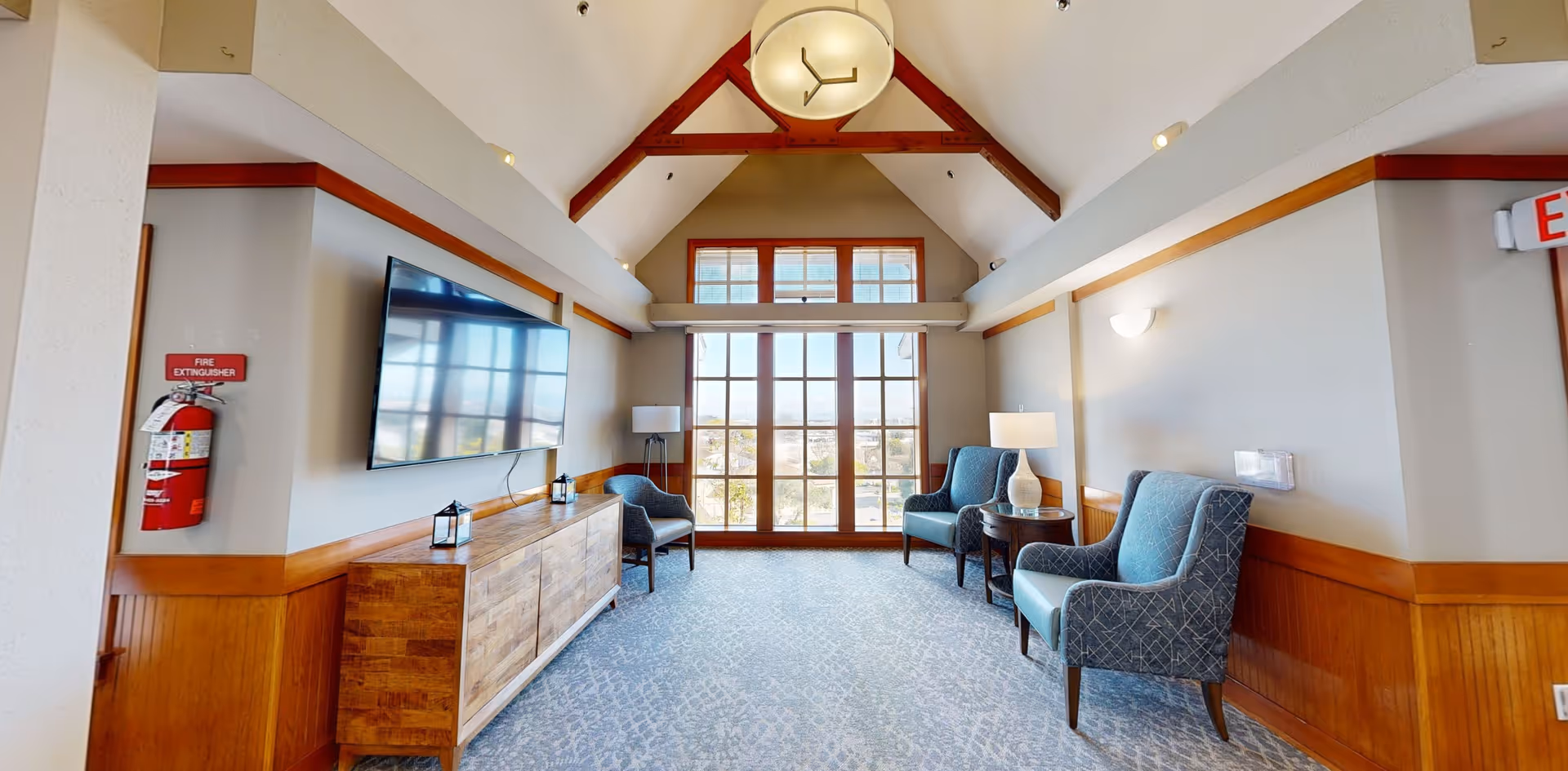 Bright communal lounge with a vaulted ceiling, large window, wall-mounted TV, wooden console, and upholstered armchairs.