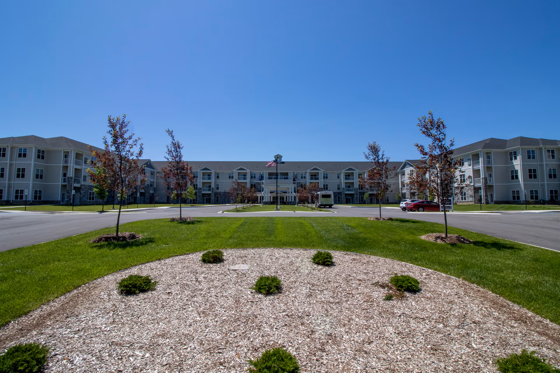 Wide exterior view of StoryPoint Chesterton, a large three-story senior living facility with beige walls and multiple windows. The building is surrounded by a well-maintained lawn, young trees, and a circular landscaped area with small shrubs. A clear blue sky is visible above.
