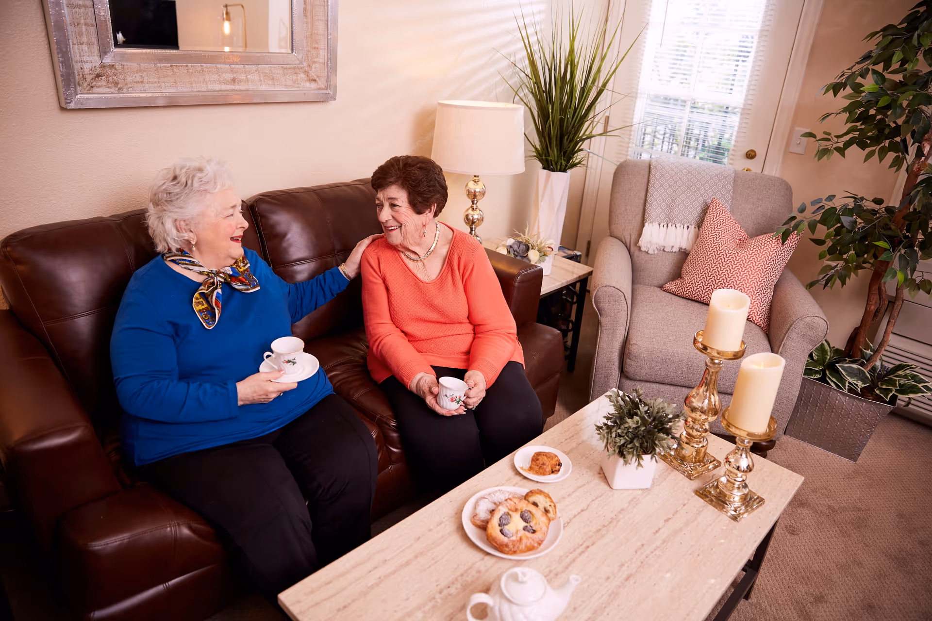 Two elderly women chat and share tea on a living room sofa.