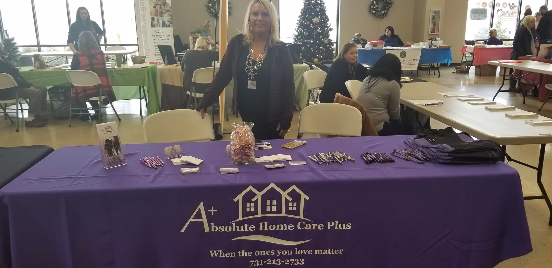 Staff members stand by a purple table promoting Absolute Home Care Plus at a community event.