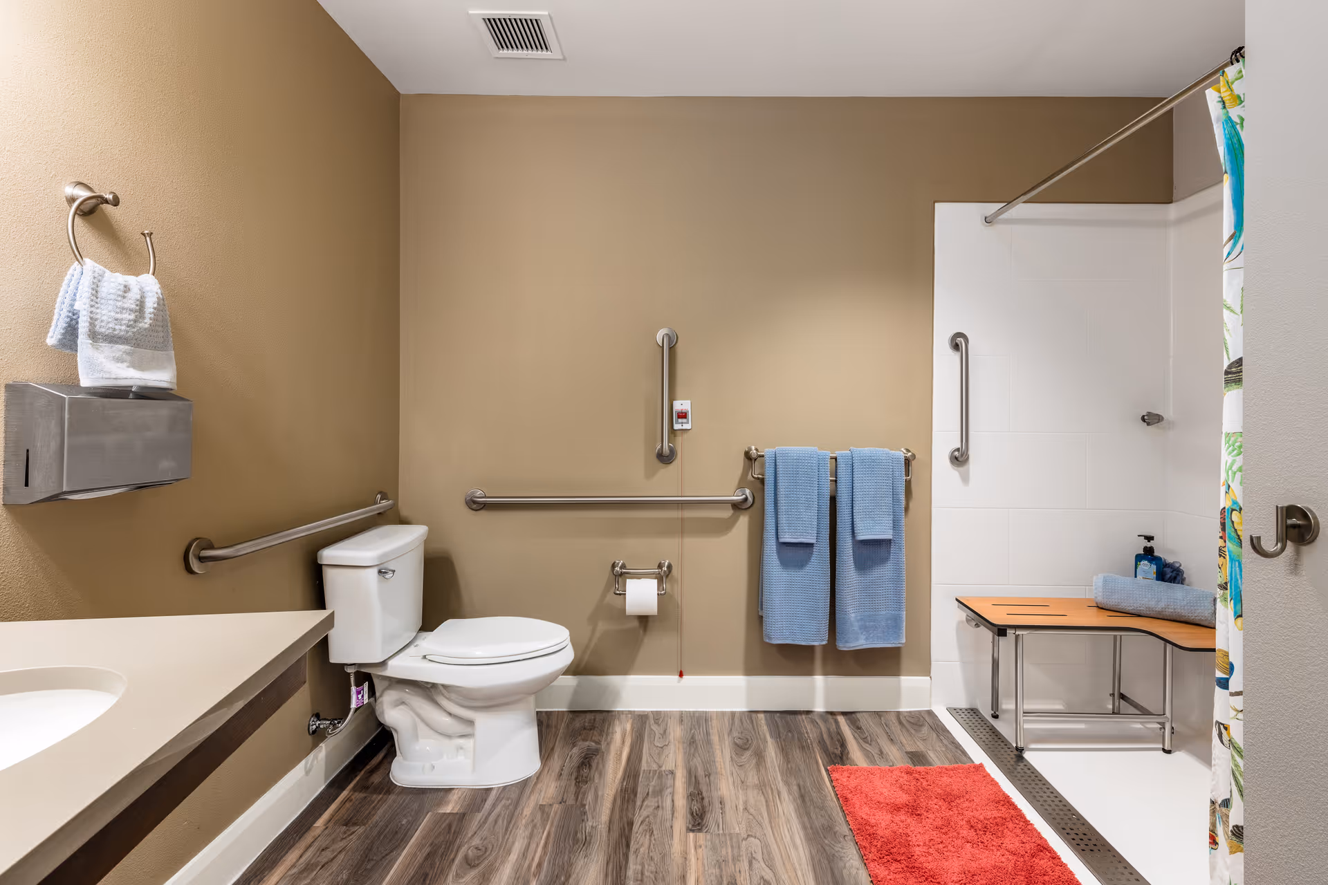 Accessible bathroom with a toilet, grab bars, towels, and a roll-in shower with a bench.