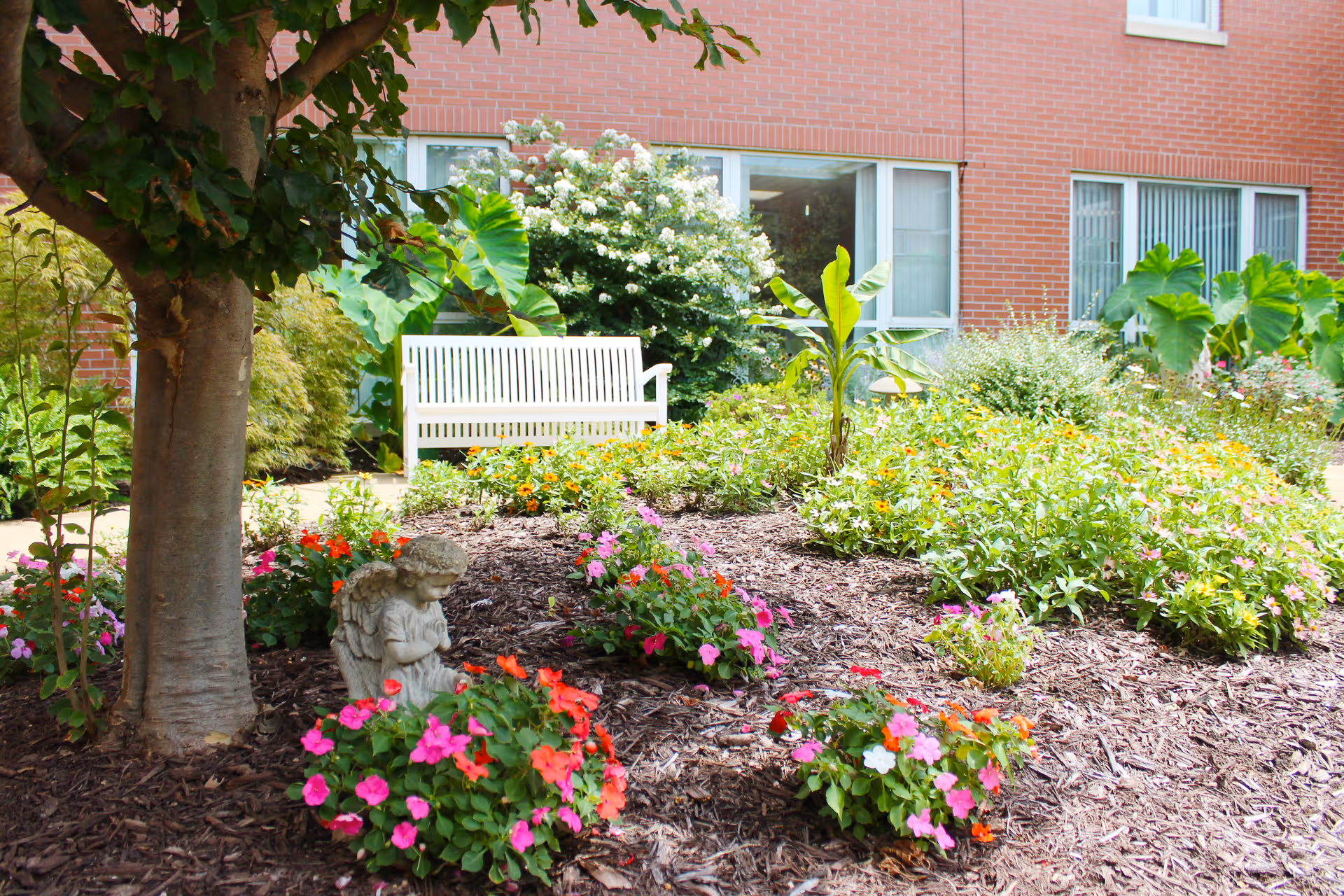 A garden area with colorful flowers and green plants surrounding a small praying angel statue. A large tree is on the left side, and a white bench is placed on a paved path near a brick building with windows in the background.