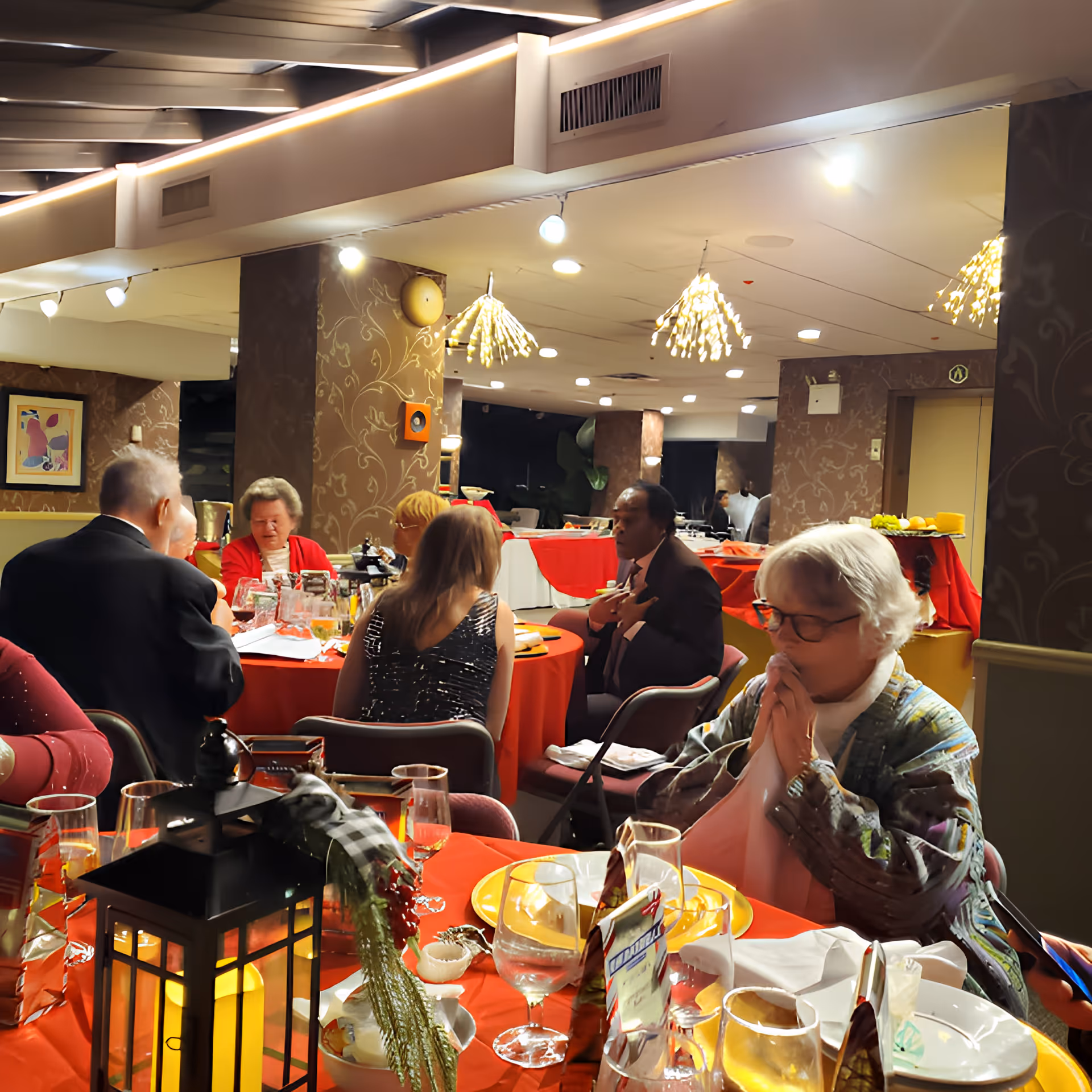 A group of elderly people seated around tables with red tablecloths in a warmly lit dining room. The tables are set with plates, glasses, and decorative items including a lantern with a candle. The room has patterned wallpaper, modern ceiling lights, and a buffet area in the background.