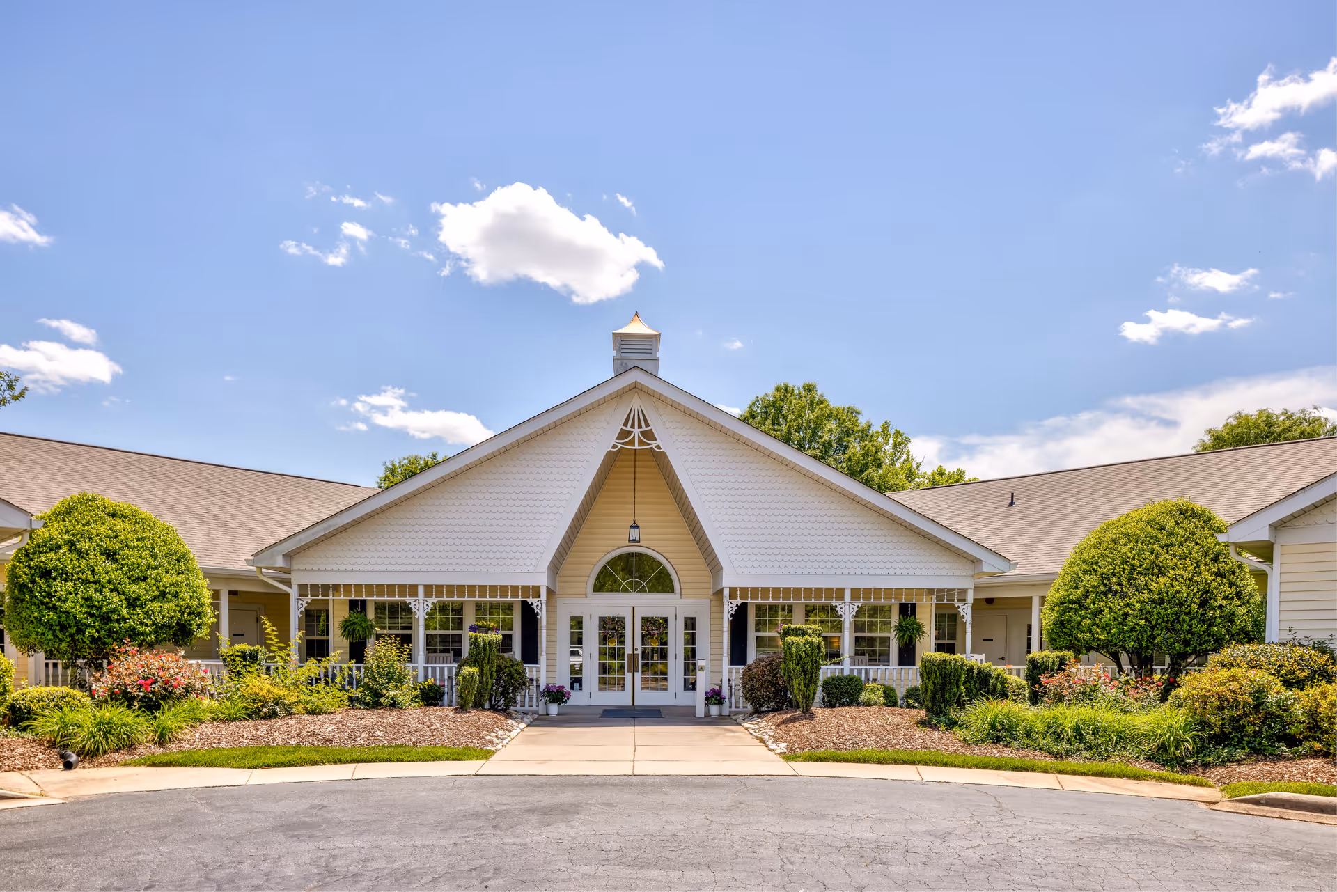 Front exterior view of TerraBella Salisbury facility with a peaked roof entrance, white siding, large windows, and well-maintained landscaping including bushes and flowers under a blue sky with some clouds.