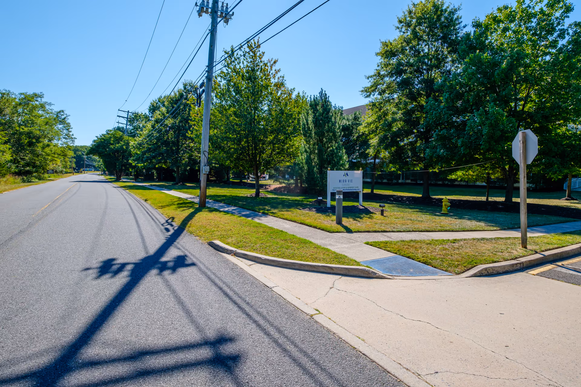 Tree-lined street and sidewalk with a grassy lawn and a small entrance sign, overhead power lines visible.