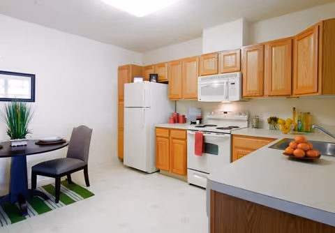 A clean kitchen with wooden cabinets, a white refrigerator, a white stove with a microwave above it, and a countertop with a bowl of oranges. To the left, there is a small dining area with a round table, two chairs, and a green-striped rug underneath. The walls are white and there is a framed picture on the wall.