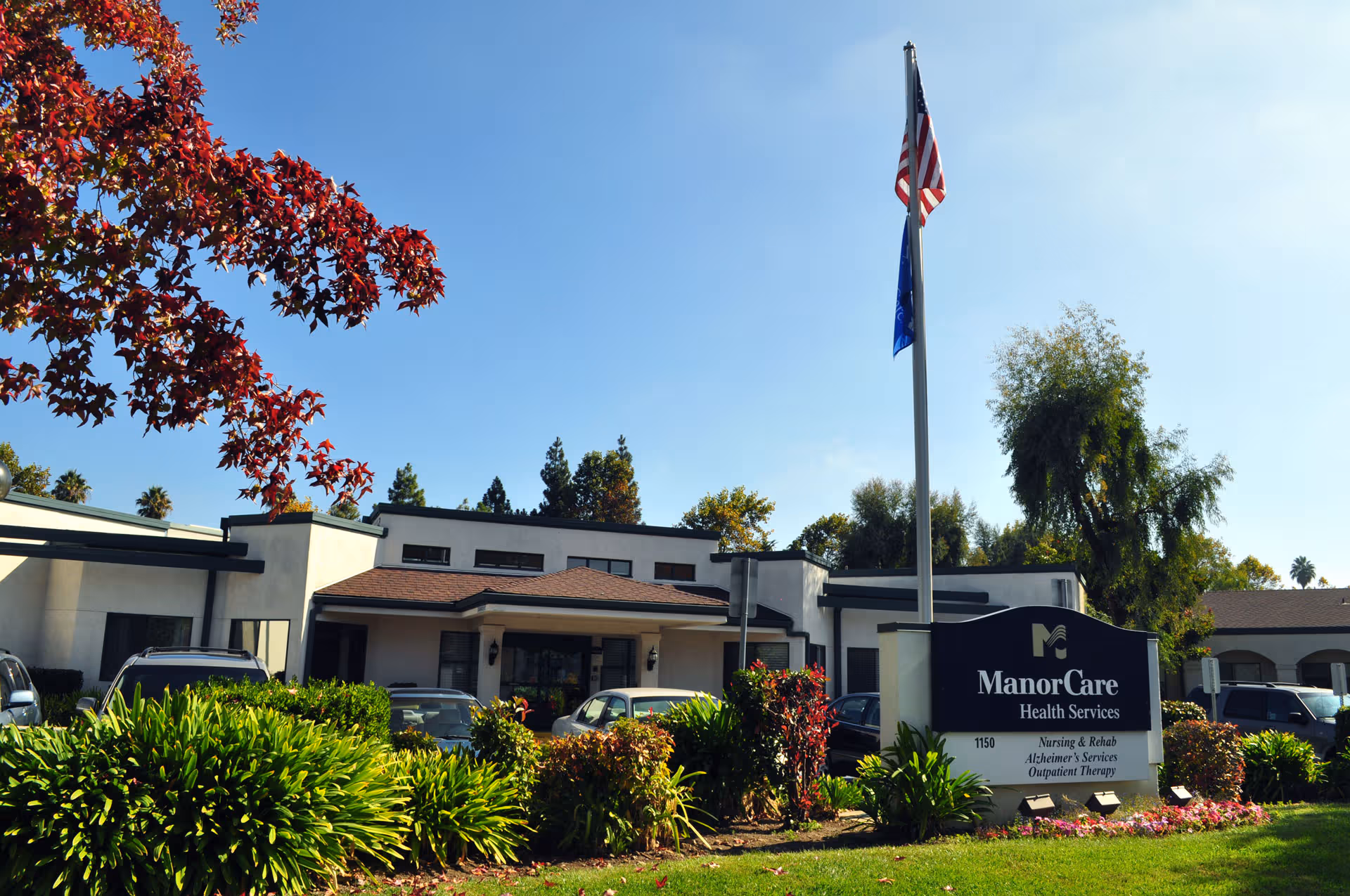 Exterior of ManorCare Health Services building with flagpole, parking, and landscaping.