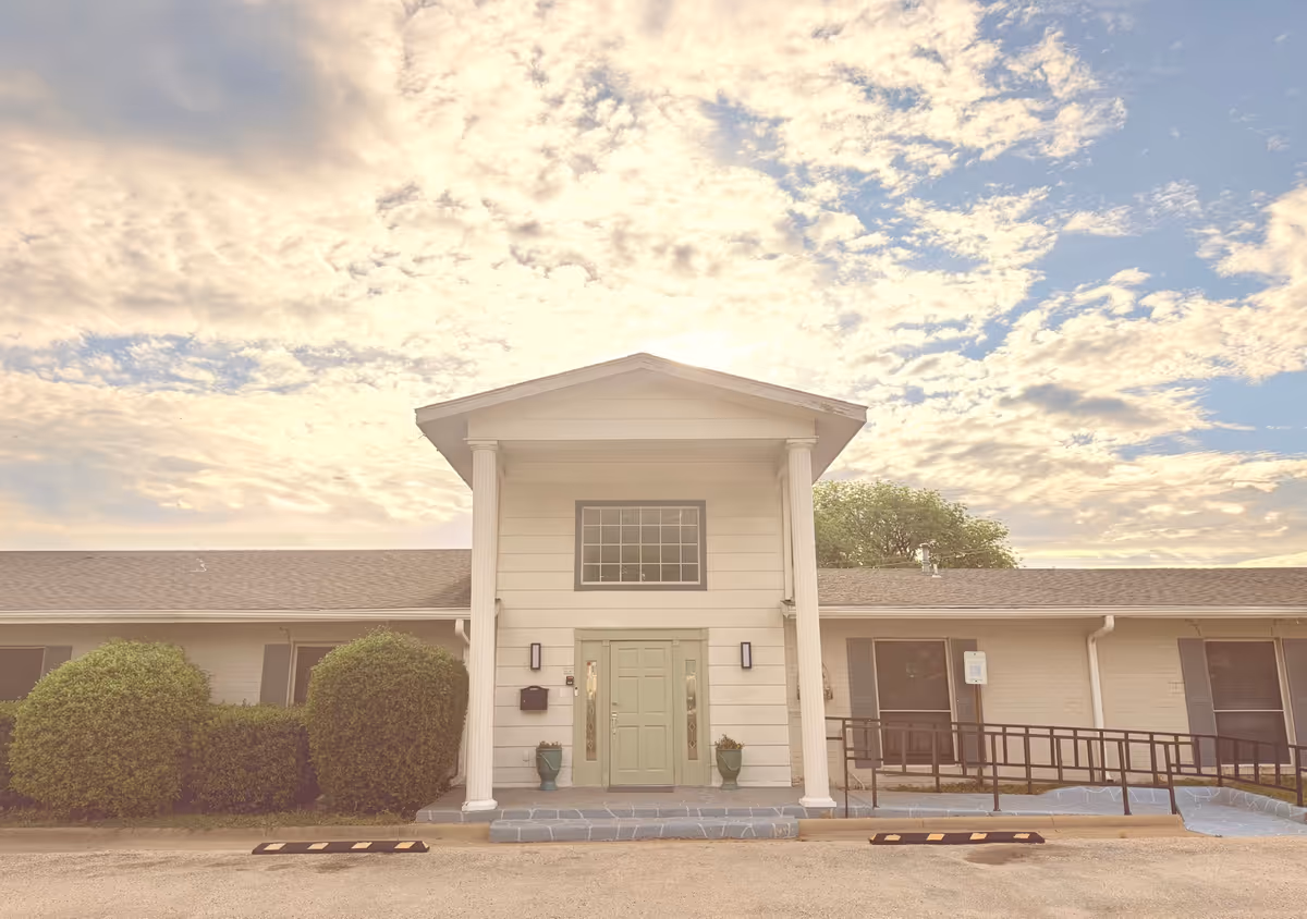 Single story senior living facility front entrance with pillars, ramp, green door, and manicured bushes.