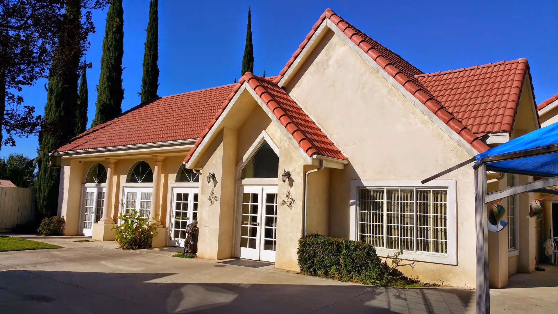 Exterior view of a building with beige stucco walls and red tiled roof under a clear blue sky. The building has multiple white-framed glass doors and windows, with some greenery and tall trees in the background. A blue canopy is partially visible on the right side.