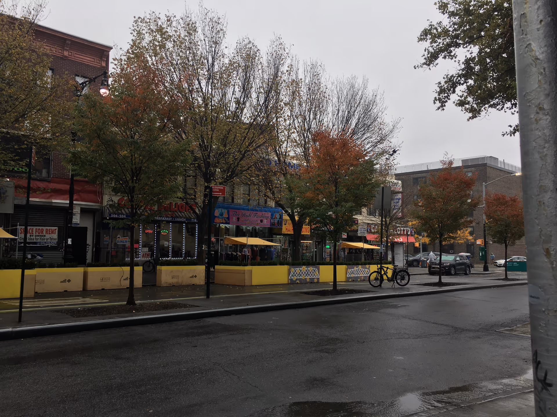 Urban street scene with shopfronts, trees, outdoor seating, and bicycles along the sidewalk.