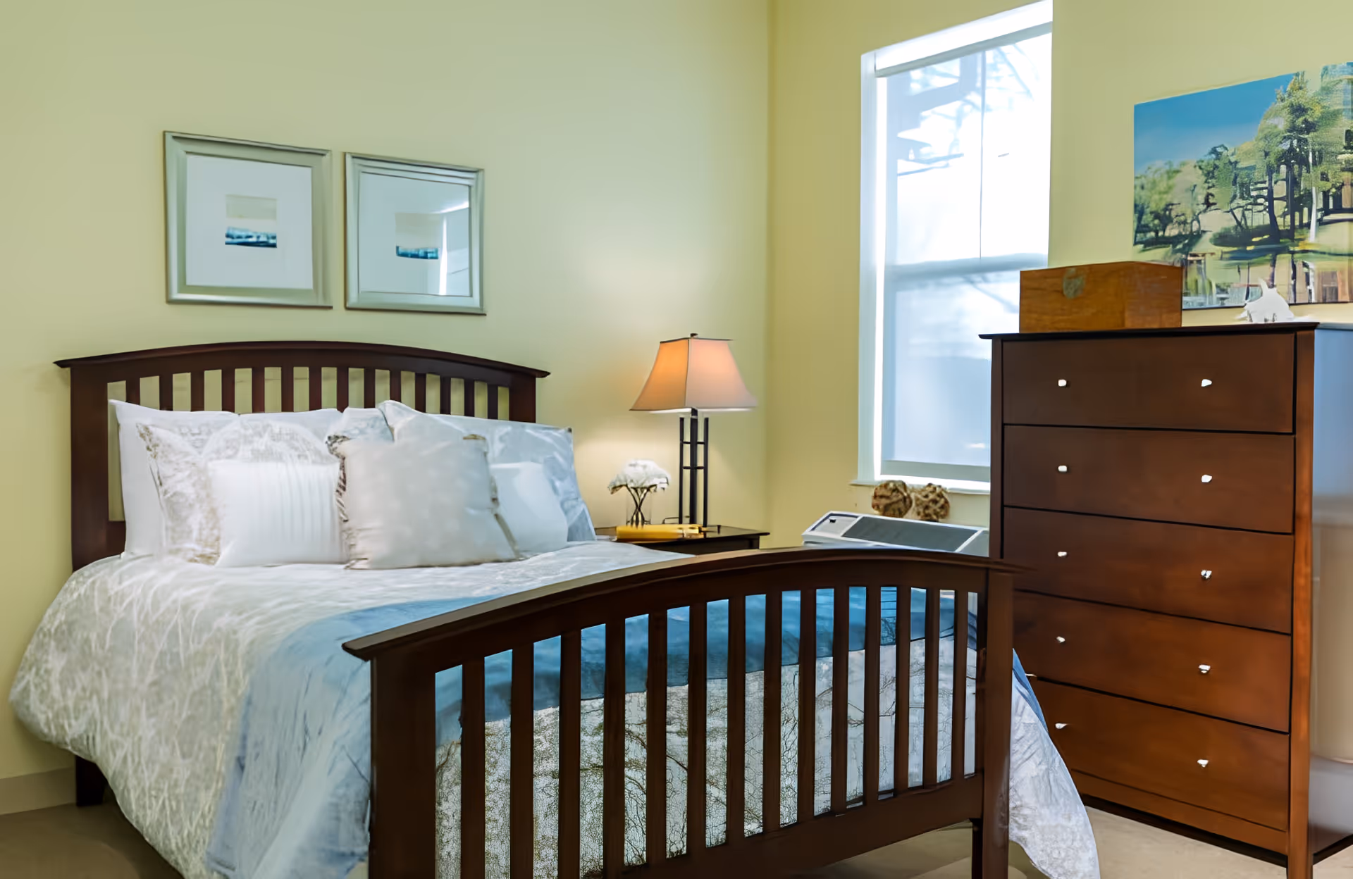 A neatly made bedroom with a wooden bed frame, bedside table and lamp, and a tall wooden dresser beneath a window.