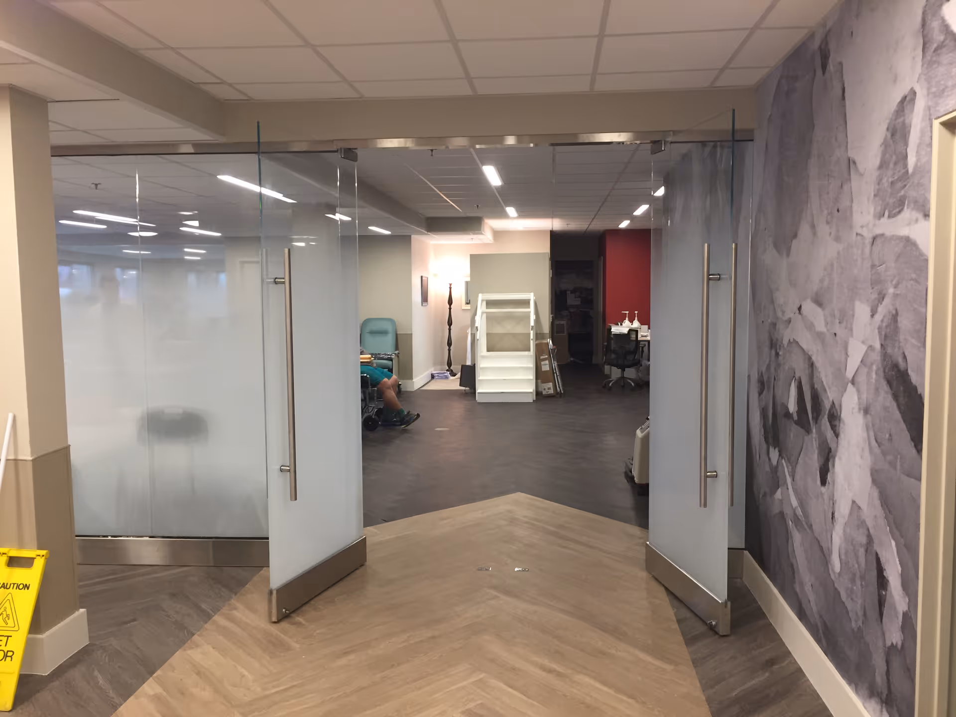 Interior view of a facility hallway with double glass doors open, showing a corridor with a person sitting in a wheelchair on the left side, a white shelving unit in the center, and a workspace with chairs and desks in the background. The floor has a wood pattern, and there is a large floral mural on the right wall.