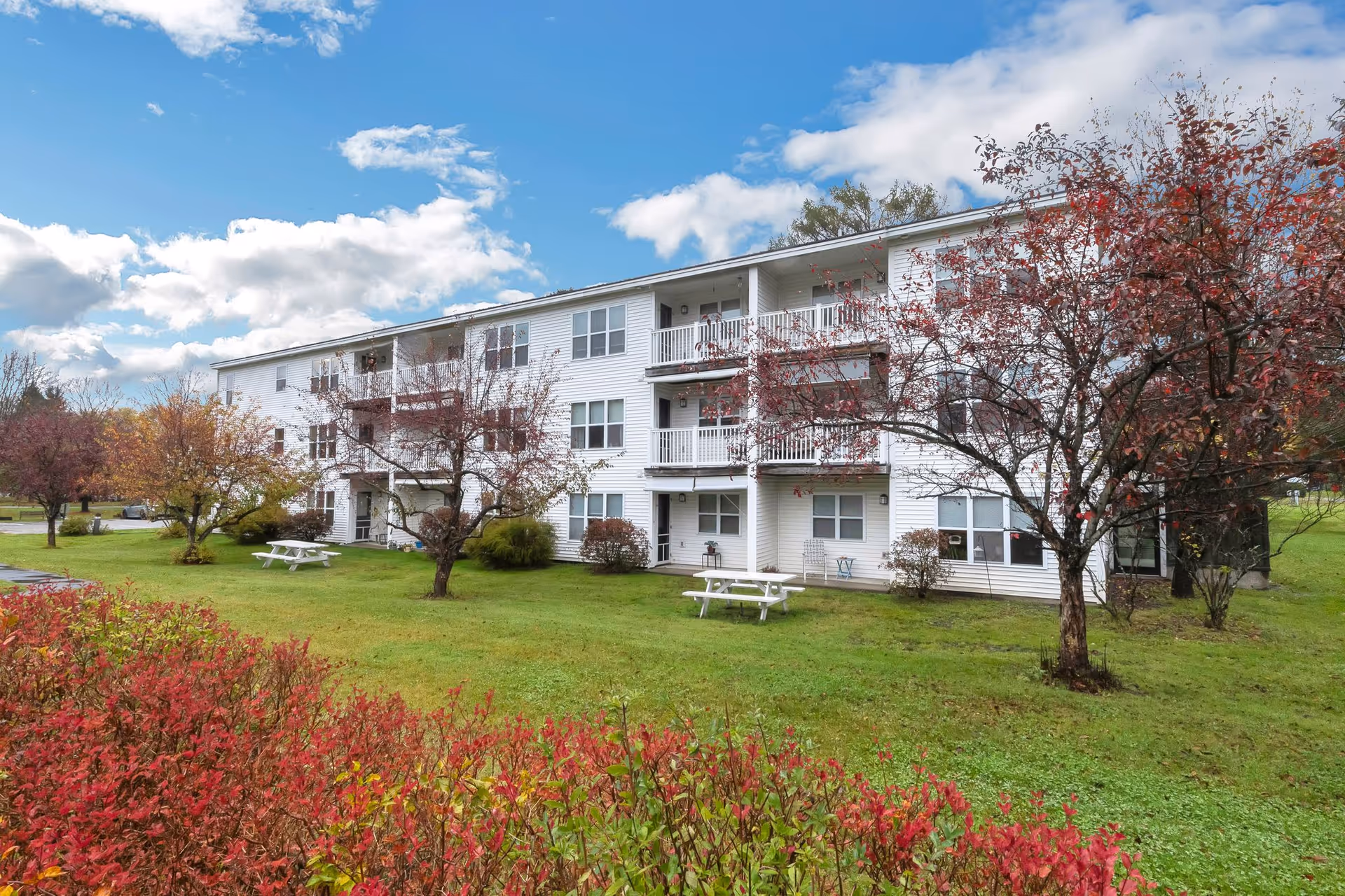 Exterior view of a three-story white senior living facility building with balconies. The building is surrounded by green grass, trees with autumn foliage, and picnic tables under a partly cloudy blue sky.